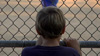 A young boy watches his friends’ baseball game through the fence.