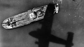 A Coronado bomber casts a shadow over a bamboo-laden Japanese boat in the water in the Western Pacific
